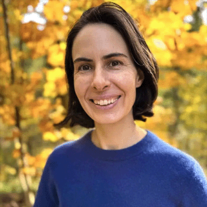A headshot of Clara Berridge wearing a blue sweater. Trees with yellow-red autumnal leaves are behind her.