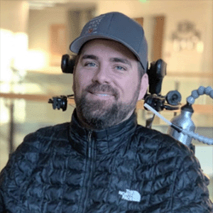 Kenny Salvini, seated in a powered wheelchair controlled by head movements with an adapted water bottle. He is wearing a puffy jacket and a baseball cap.