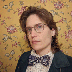 Headshot of Rua Williams. They look somberly at the camera and wear a floral bowtie with a floral shirt and blazer.