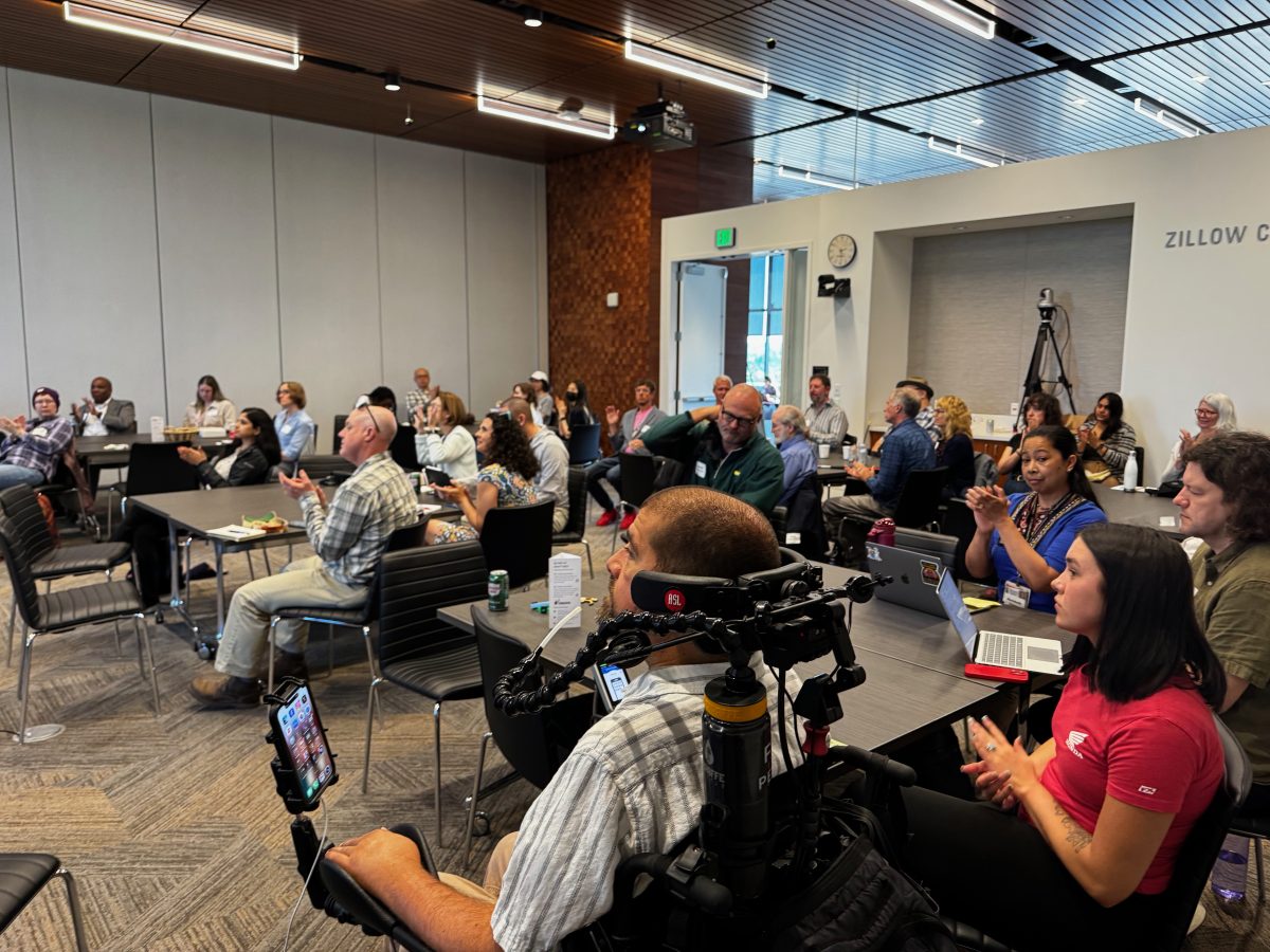 Photo of the audience. About 25 people are clapping, seated around tables in a large, well lighted room.