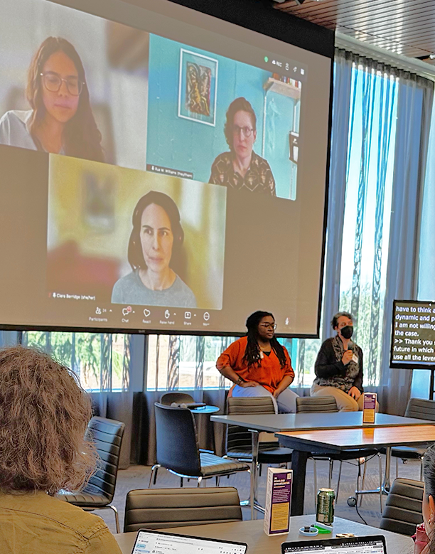 Photo from the Examining AI through a Disability Perspective panel. Panelists Jazette Johnson and Jen Mankoff are seated at the front of the room. Behind them on a large Zoom screen are Ariana Aboulafia, Clara Berridge, and Rua Williams. A monitor next to Mankoff displays auto captions.