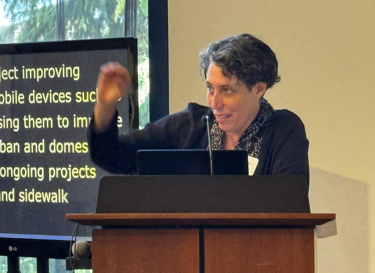 Jennifer Mankoff at the podium, speaking and gesturing. A monitor behind her displays auto captions.