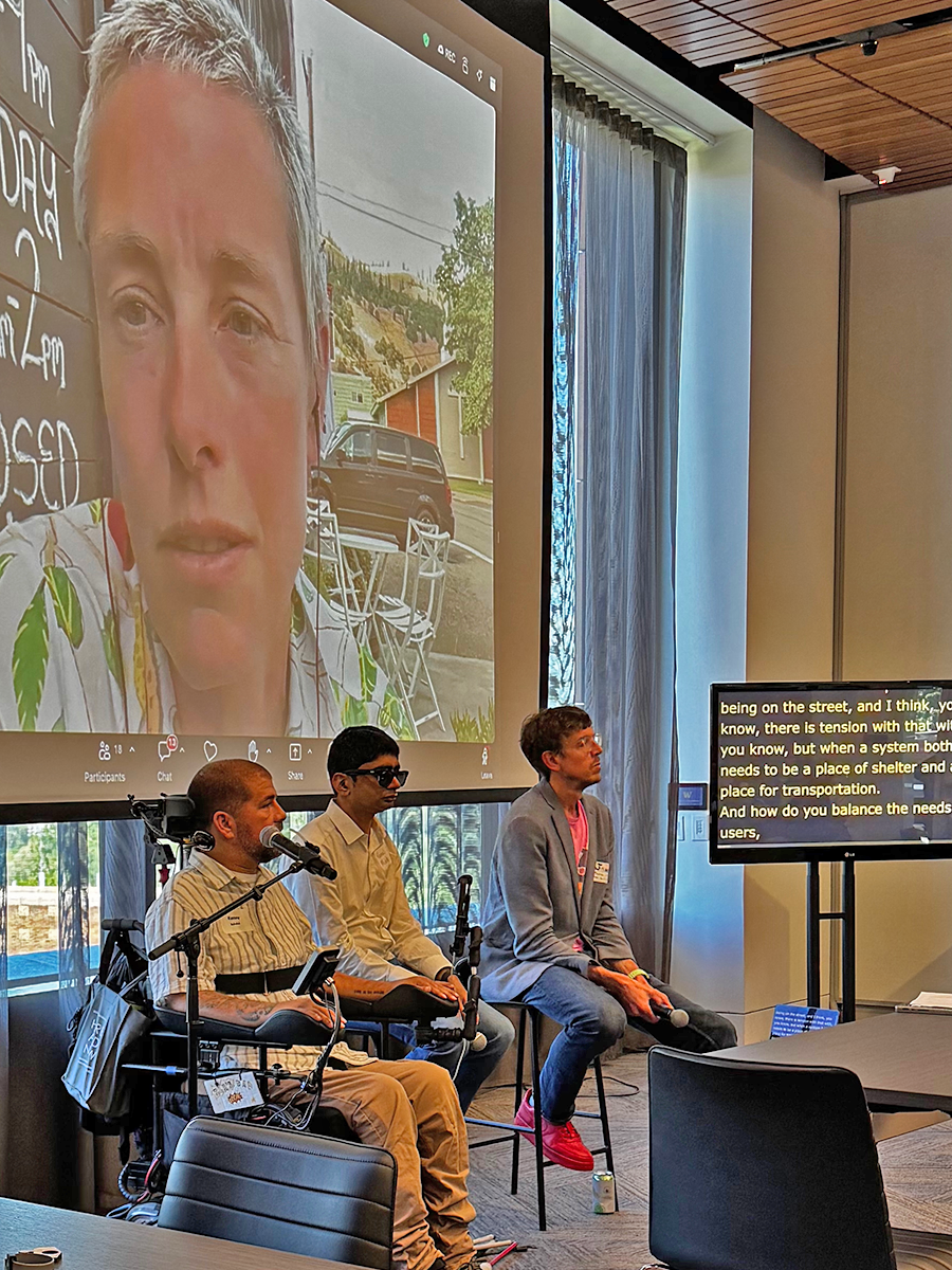 Photo from the Transportation and Urban Mobility panel. Panelists Kenny Salvini and Kunal Mehta are seated at the front of the room along with moderator Jon Froehlich. Behind them on a large Zoom screen is panelist Anna Zivarts. A monitor next to Froehlich displays auto captions.