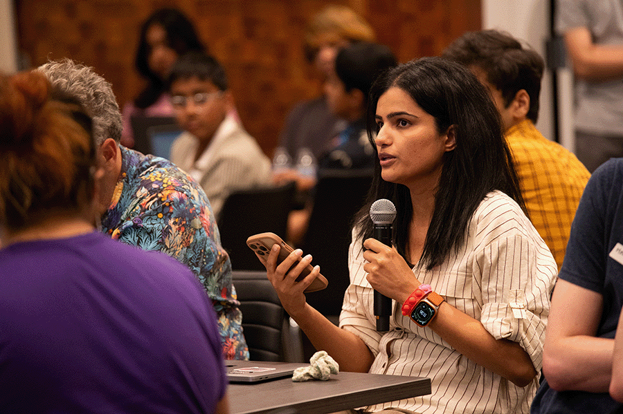 A participant seated in the audience speaks while holding a microphone in one hand and a mobil phone in the other. Behind her are nine more participants at CREATE Accessibility Hackfest - Summer 2026.