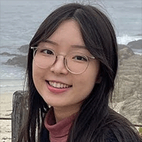 A young woman with long dark hair. Behind her is a rocky beach and waves.