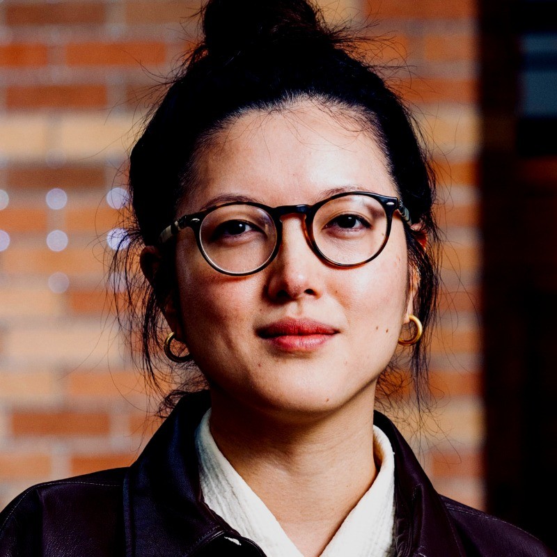 Stacy Hsueh, an Asian woman. She wears a white collared shirt and black blazer. In the background is a brick wall.