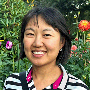Lucy Lu Wang, a smiling East Asian woman with chin-length black hair, wearing a black and white striped shirt. Behind her are trees and dahlias.