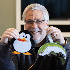 Paul Dietz, a man with silver hair and bear, holding up two paper animatronic puppets: a talking penguin and a talking salmon.