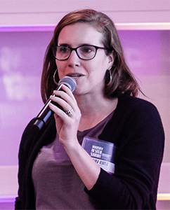 Amy Hurst, a white woman with brown hair and glasses, holding a microphone and talking in a purple room.