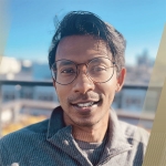 Headshot of Ather Sharif outside on a sunny balcony with blue sky behind him