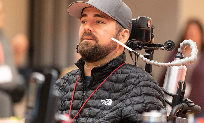 Kenny Salvini, seated in a powered wheelchair controlled by head movements with an adapted water bottle. He is wearing a puffy jacket and a baseball cap.