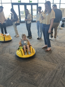 Attendees watch a toddler demonstrate a ride-on toy adapted for mobility.