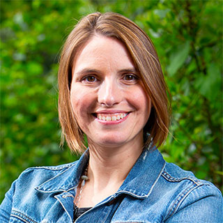 Julie Kientz, a white woman with medium-long brown hair and brown eyes. She is smiling, wearing a denim jacket with arms crossed, in front of a green hedge