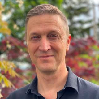 Mark Harniss a white man in his 50s with short brown hair and blue eyes, standing in front of fall-colored leaves.