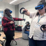 Two student participants at the 2019 OurCS workshop use virtual reality goggles and hand controllers