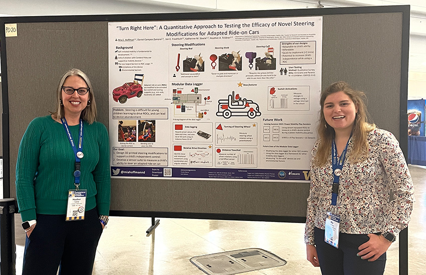 Heather Feldner and Mia Hoffman stand next to their poster board about adapted ride-on cars research at a conference.