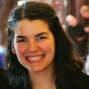 Sarah Coppola, a white woman with long, black curly hair. She is smiling at the camera and the background is blurred.