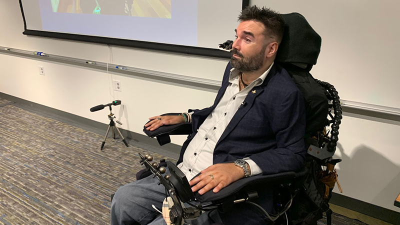 Tyler Schrenk making a presentation at the head of a lecture room. He has brown spiky hair, a full beard, and is seated in his power wheelchair.