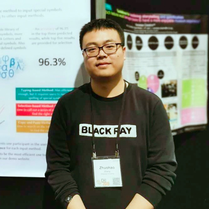 Zhuohao (Jerry) Zhang standing in front of a poster, wearing a black sweater and a pair of black glasses, smiling. 