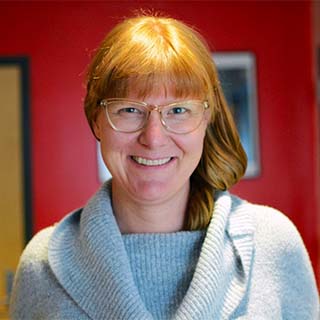 Image of Abigale Stangl, a woman with medium length ginger/red hair smiling and wearing a sweater clear framed glasses She is standing in an academic lab, with brightly colored walls