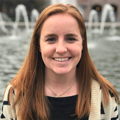 Alyssa Spomer, a young woman with long red hair, smiling and standing in front of Drumheller Fountain on the UW Seattle campus.