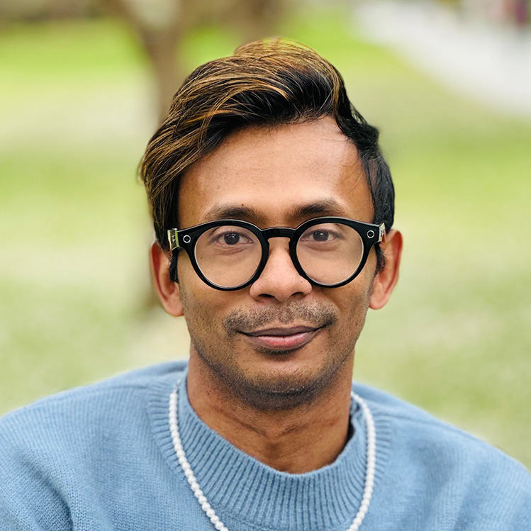 Headshot of Ather Sharif, a brown man with black and brown hair smiling at the camera, wearing eyeglasses, a light blue sweater, and a pearl necklace. The background is blurred.