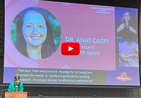 Olivia Quesada stands at a podium to accept the 2023 Human Rights Educator Award for Anat Caspi whose photo is shown on a large screen in the background.