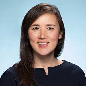 Headshot of Emily Whiting, a mixed race woman with shoulder-length brown hair, smiling at the camera. She is in front of a light blue background.