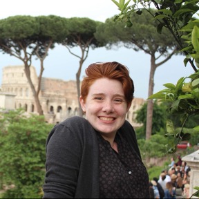 Emma McDonnell, a white woman in her 20s with short red hair, freckles, and a warm smile. in the background: a lush landscape and the Colosseum. 