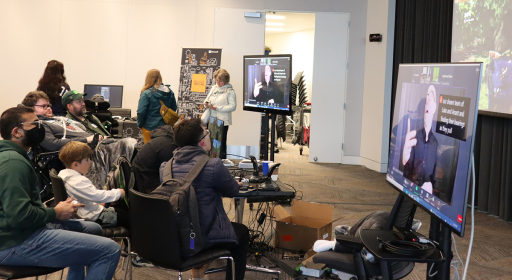A group of gamers - young and less young, some in wheelchairs or using adaptive devices - play a game shown on a large screen while an ASL interpreter is shown on a secondary screen.