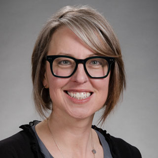 Headshot of Heather Feldner, smiling brightly. She is a white woman with short brown and grey hair, and wears dark rimmed glasses, a gray shirt and black sweater.