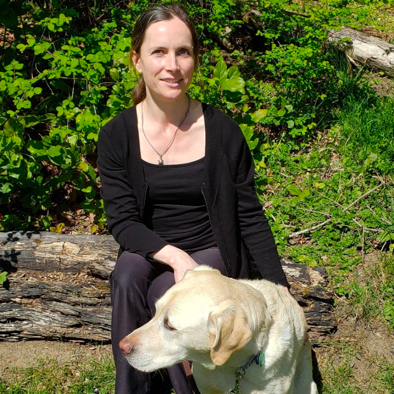Natalie McCarthy, a white woman with long, brown hair, sitting on a log with her guide dog, Vidal.