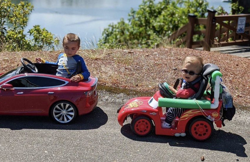 Two children ride in small toy cars, one of which has an adapted steering wheel to make it accessible for the child to use.