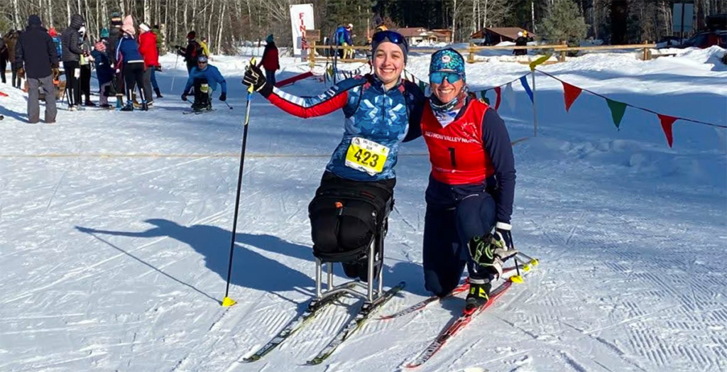 Nicole Zaino, wearing ski clothes and a racer’s bib, poses on a snowy ski trail with BethAnn Chamberlain of U.S. Paralympics Nordic Skiing.